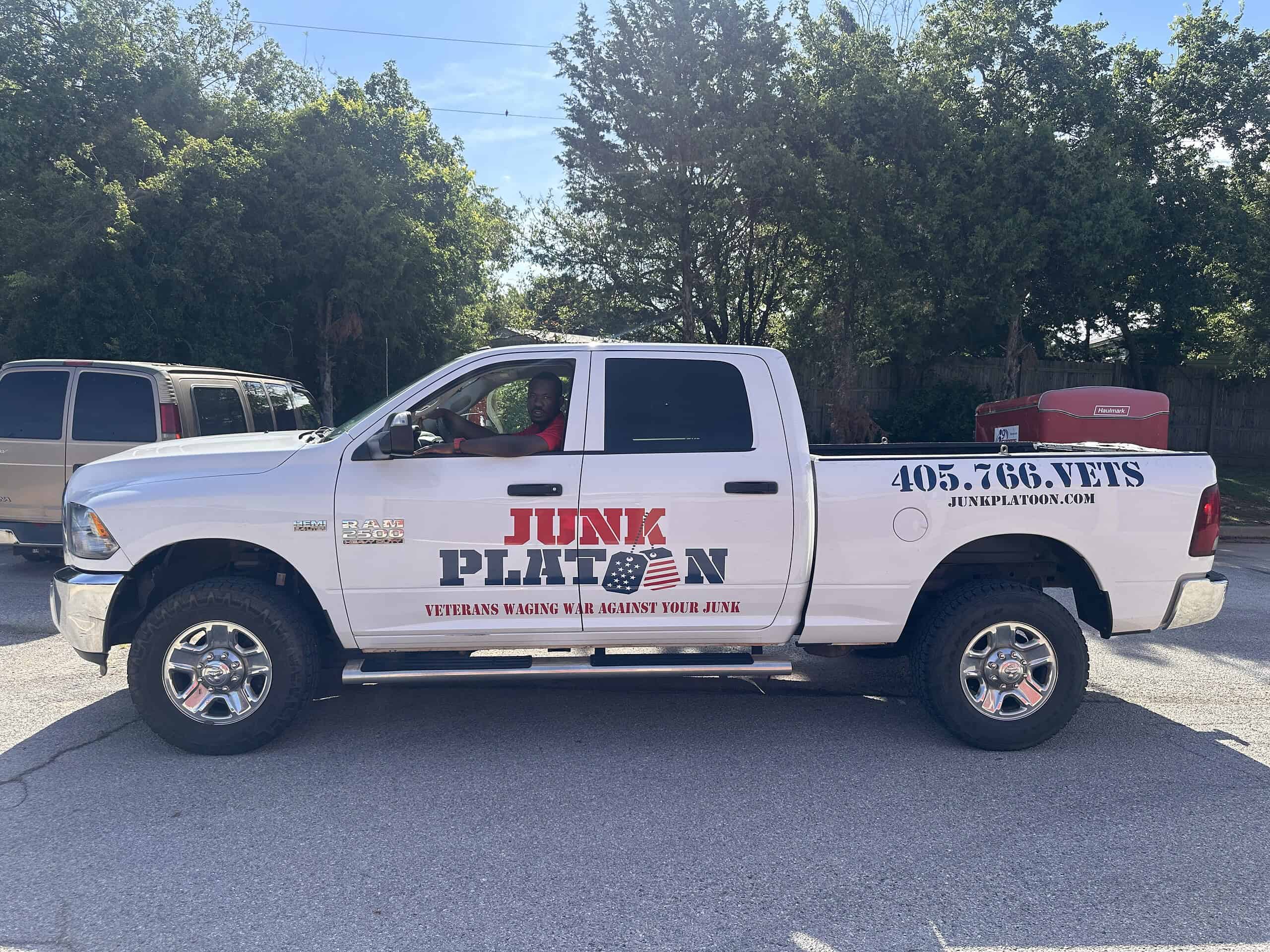 Junk Platoon truck with veteran logo, military junk removal service. Alternative Text: White Junk Platoon truck with veterans service branding parked outdoors.